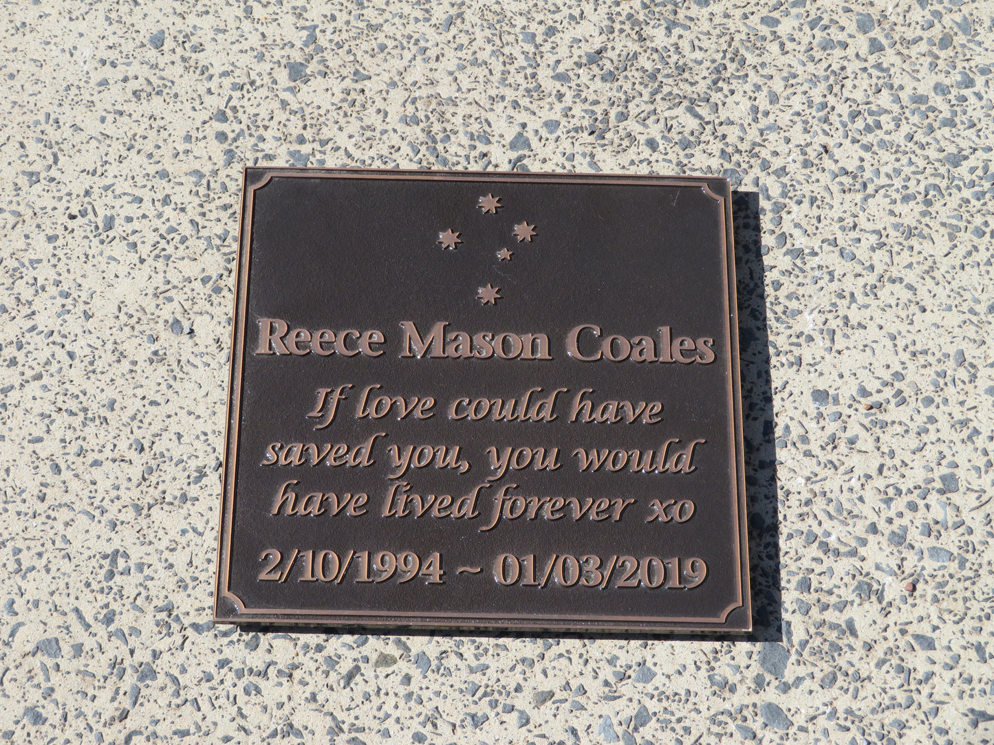 large square bronze plaque with memorial inscription for a person. It is placed on a concrete floor