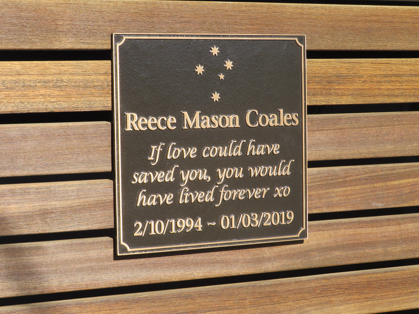 large square bronze plaque with memorial inscription for a person. It is placed on planks of wood.