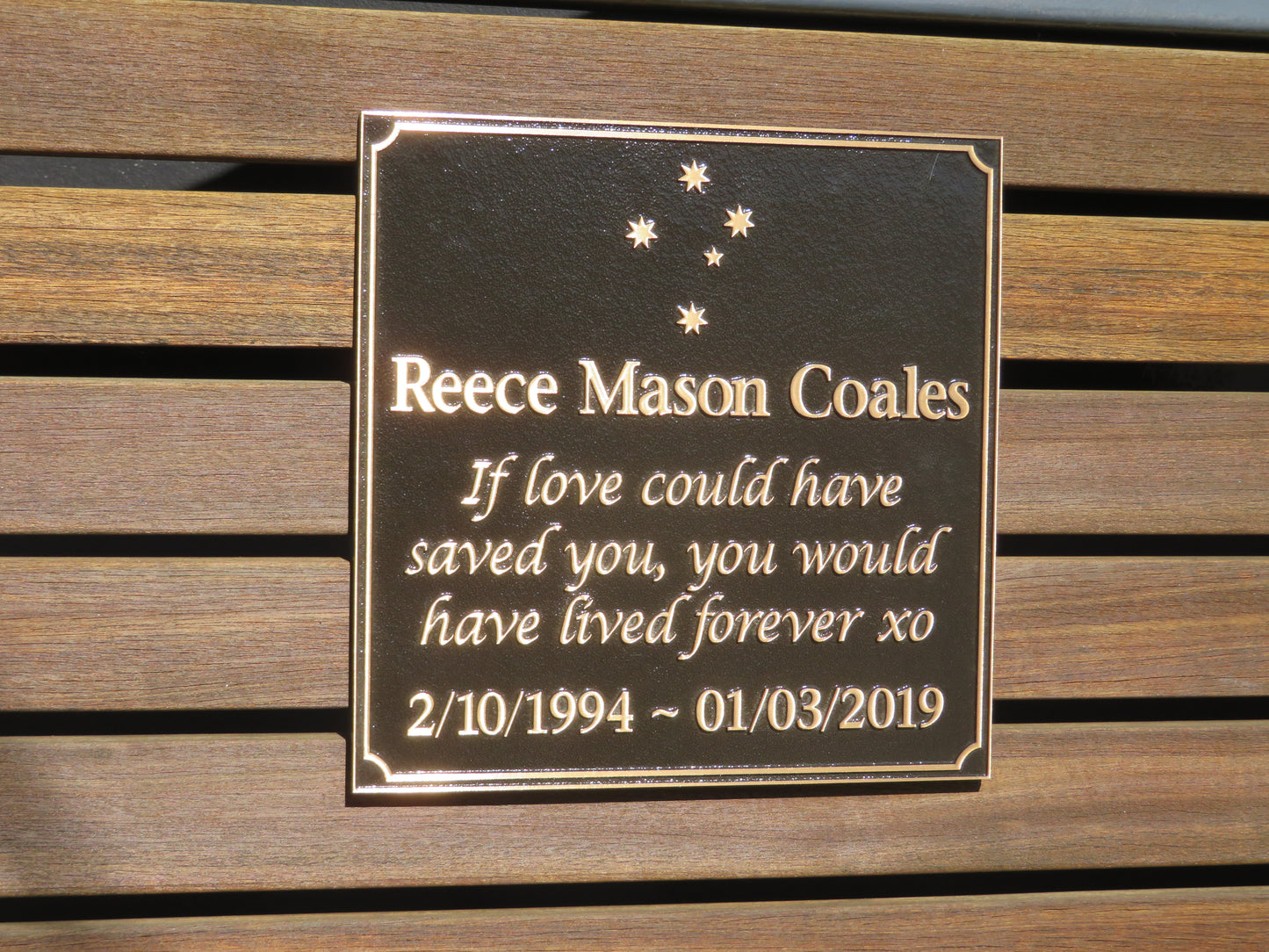 large square bronze plaque with memorial inscription for a person. It is placed on planks of wood.