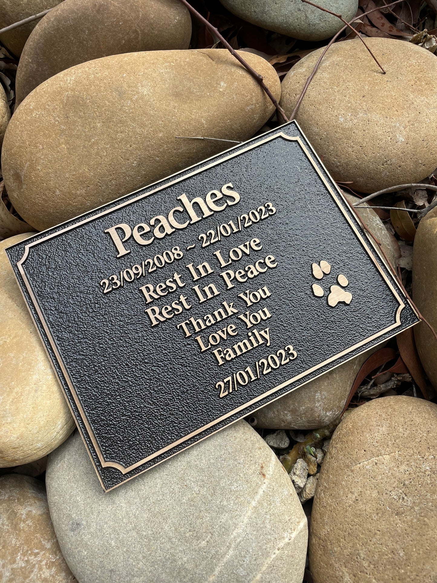 rectangle bronze plaque with memorial inscription for a pet with a custom paw print. The plaque is placed on a bed of riverstones